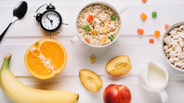 Oatmeal porridge, fruits, cereals and milk with alarm clock on white table