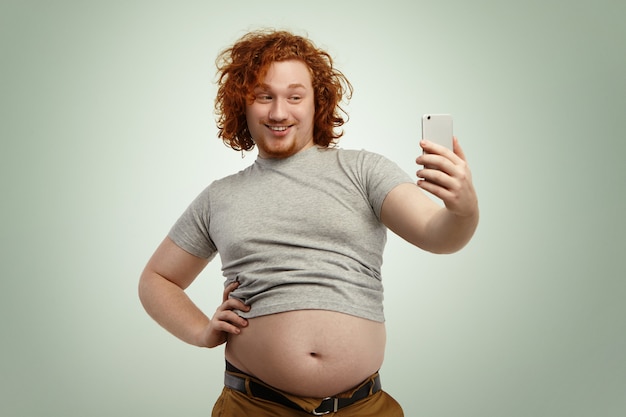 Foto gratuita obeso giovane maschio con i capelli ricci di zenzero e la barba che tiene il telefono cellulare, in posa per selfie, guardando con un sorriso civettuolo mentre la sua pancia grassa pende dalla t-shirt grigia e pantaloni jeans stretti