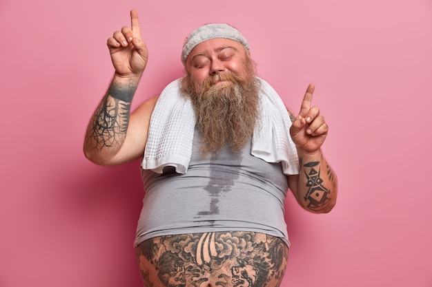 Obesity and sport concept. Joyful overweight man dances carefree has sweaty body tattooed arms points upwards isolated on pink wall, does exercises at home, burns calories after eating fast food