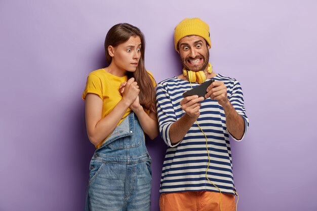 Obsessed couple play video game on smartphone, look nervously at display, eager to win, wear fashionable clothes, have worried excited expressions, isolated over purple wall. Youth, addiction