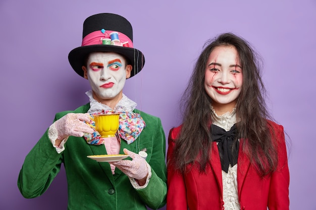 October holiday. Serious dissatisfied hatter drinks tea poses near cheerful brunette woman with skull makeup