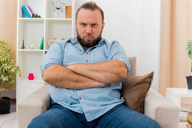 Offended adult slavic man sits on armchair crossing arms inside designed living room