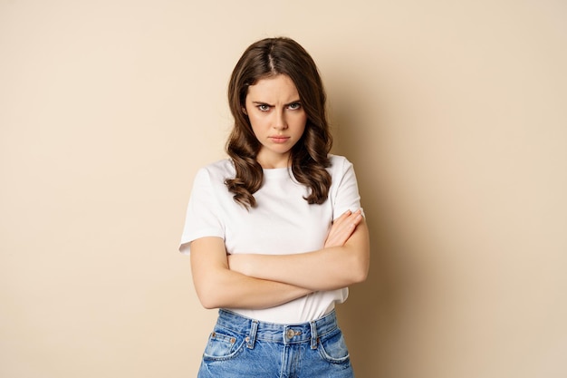 Offended angry woman cross hands on chest, frowning and sulking insulted, standing over beige background