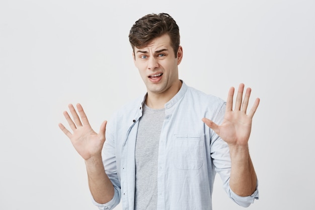 Offended caucasian young male has rush reaction on what he heard, raises hands, like saying stop that, has displeased and angry expression, isolated .