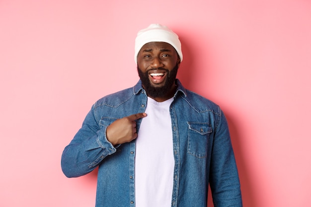 Offended and confused african-american man pointing at himself, staring at camera bothered, being accused, standing over pink background.