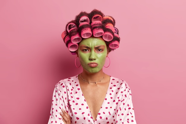 Offended displeased young woman applies beauty green face mask, tired of waiting for cosmetic product effect, wears curlers on hair, poses against rosy . Housewife makes hairstyle.