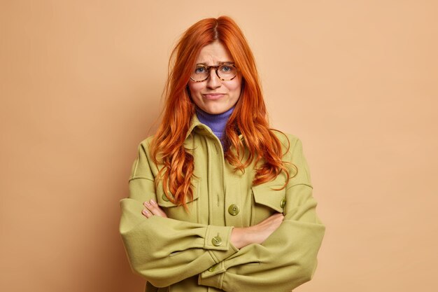 Offended dissatisfied redhead young woman keeps arms folded waits for explanations from boyfriend has hurt feelings dressed in fashionable green jacket smirks face.