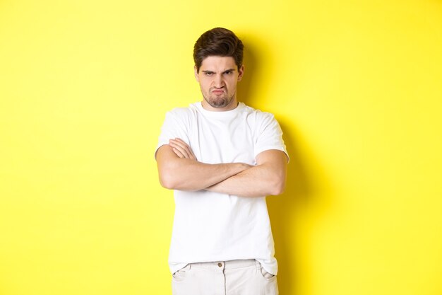 Offended and mad guy, frowning and sulking, holding hands crossed on chest, standing angry against yellow background