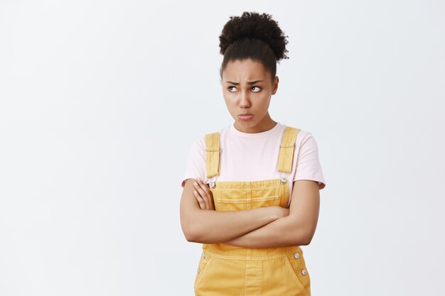 Offended upset and jealous young female student with dark skin, frowning and sulking, pursing lips, staring up while feeling insulted, holding hands crossed on chest from anger and disappointment