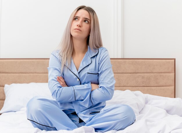 Offended young beautiful woman in blue pajamas sitting on bed looking up with hands crossed on chest in bedroom interior on light background