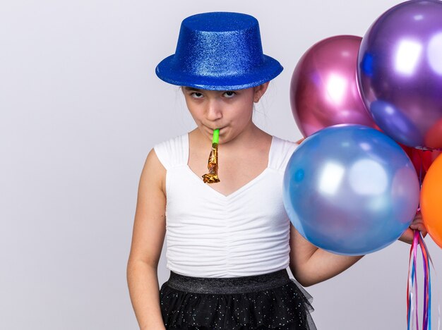 offended young caucasian girl with blue party hat holding helium balloons and blowing party whistle isolated on white wall with copy space