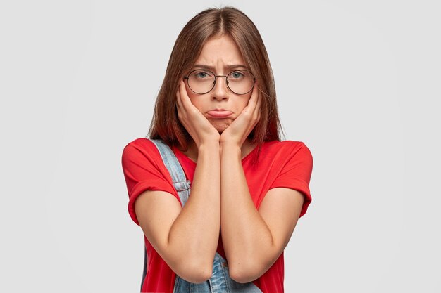 Offended young cute brunette female has abused discontent expression, purses lower lip, keeps hands on cheeks, wears round spectacles and red t-shirt, doesn't want to work or study, poses indoor