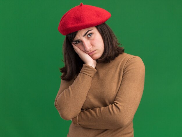 Free photo offended young pretty caucasian girl with beret hat puts head on hand isolated on green wall with copy space