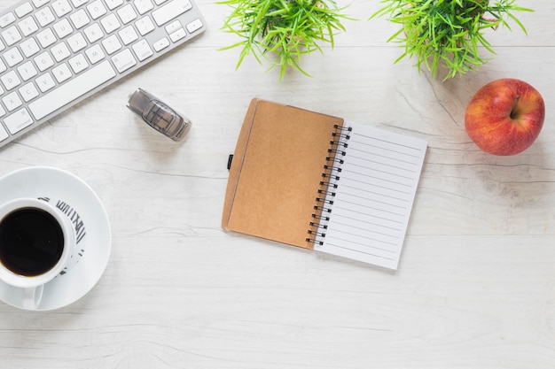 An office desk with diary; coffee cup; apple; stapler and keyboard on wooden desk