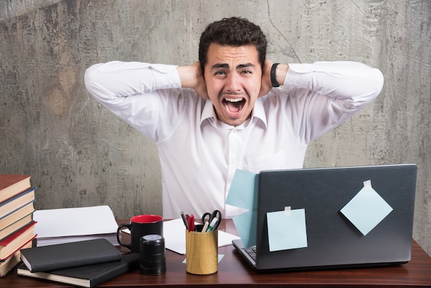 Free photo office employee screaming while holding his ears at the office desk.