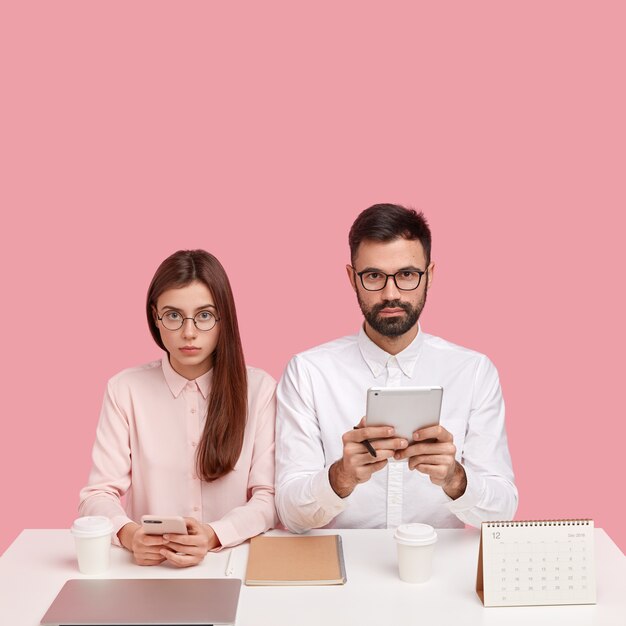 Office perfectionism concept. Serious colleagues, young workers in white elegant clothes, use modern technologies, pose at desk, drink takeaway coffee, isolated over pink wall, check newsfeed