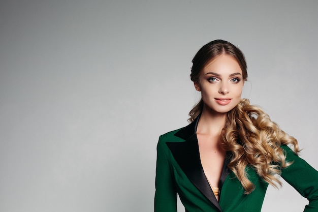 Stile rigoroso da ufficio ragazza bionda con bei capelli ondulati in una giacca da ufficio verde elegante sorridente amichevole con moderazione guarda la telecamera isolare su uno sfondo grigio