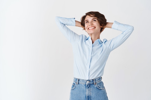 Office woman dreaming of vacation, looking relaxed up and smiling, holding hands behind head and resting from work, standing on white wall