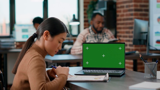 Office worker analyzing charts on paper and looking at greenscreen on laptop, using chromakey mockup template and isolated copyspace with blank background. Woman with disability.