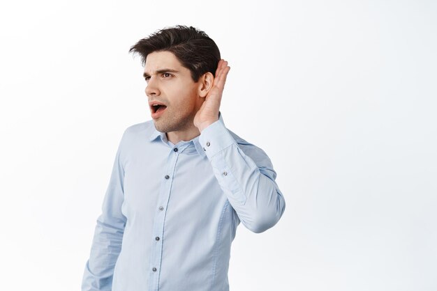 Офисный работник подслушивает, держась за руку возле уха и слушая поближе, подслушивает слухи на работе, стоя на белом фоне