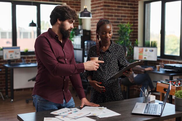 Office worker pointing out wrong accounting data while expectant mother having a clipboard in hand. Marketing company employees reviewing profitable strategy ideas and project startup partners.