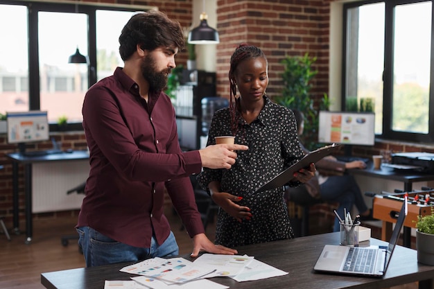 Office worker pointing out wrong accounting data while expectant mother having a clipboard in hand. Marketing company employees reviewing profitable strategy ideas and project startup partners.