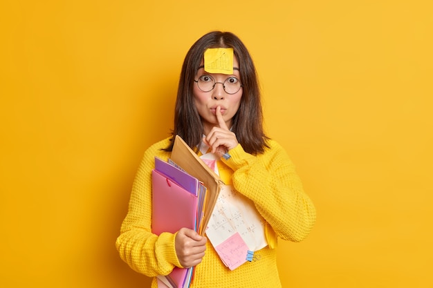 Office worker with sticker on forehead makes silence gesture holds folders wears round spectacles and jumper.