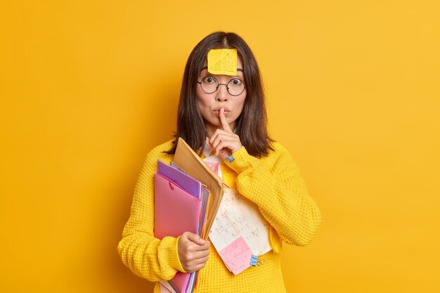 Office worker with sticker on forehead makes silence gesture holds folders wears round spectacles and jumper.
