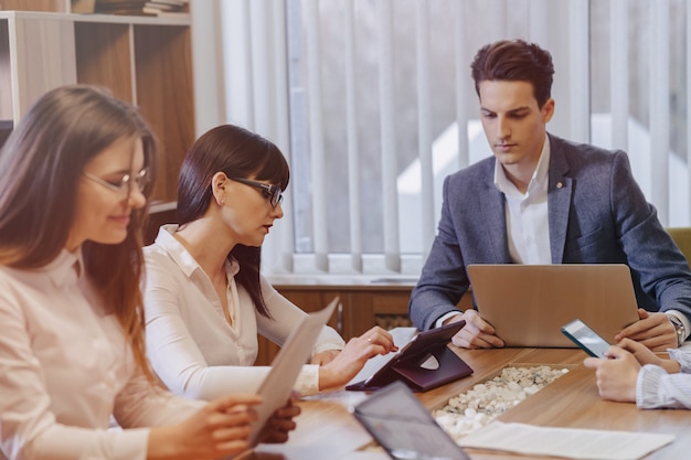 Office workers hold a meeting at one desk for laptops, tablets and papers, on the background a large TV set on a wooden wall