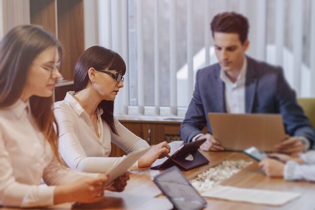 Office workers hold a meeting at one desk for laptops, tablets and papers,  a large TV set on a wooden wall