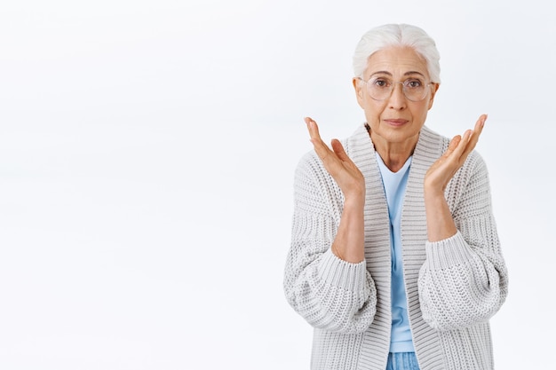 Oh dear grandmother looking at child grown up so fast, raising hands near cheeks from cuteness and lovely scene, being touched and impressed, smiling happily, standing white wall in glasses