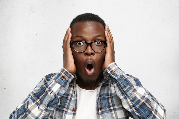 Free photo oh my god. human facial expressions, emotions and feelings. bug-eyed stylish young dark-skinned man in spectacles exclaiming in shock, keeping hands on head, astonished with some unexpected news