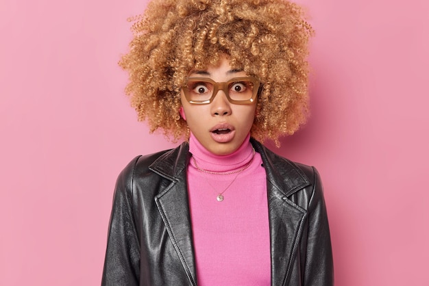 세상에. 놀란 곱슬머리의 젊은 유럽 여성은 분홍색 배경에 격리된 터틀넥 가죽 재킷을 입은 카메라를 쳐다보는 믿을 수 없는 뉴스를 듣고 입을 벌리고 있습니다. 와 대단하다