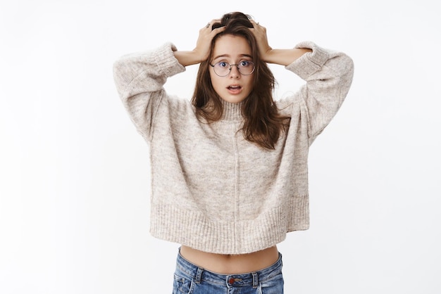 Oh my what to do Silly and clueless goodlooking confused girl in glasses and sweater holding hands on head open mouth from surprise unknowing how solve situation standing puzzled and clueless