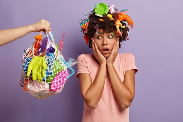 Oh no, do not pollute nature with plastic waste! Unhappy ethnic woman looks with shocked expression at bag full of plastic garbage, cleans planet, poses indoor. Earth Day and volunteering concept