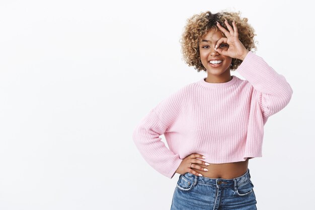 Oh yes, that is it. Delighted carefree bright optimistic dark-skinned female with blond hair in trendy sweater standing confident, enthusiastic showing okay gesture on eye and looking through ok sign