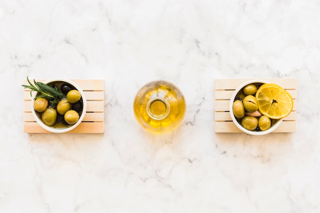 Oil bottle between the two bowls of olive with rosemary and lemon slice