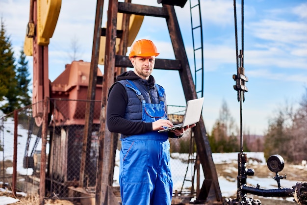 Oil engineer with a laptop standing next to an oil rig making notes in his computer