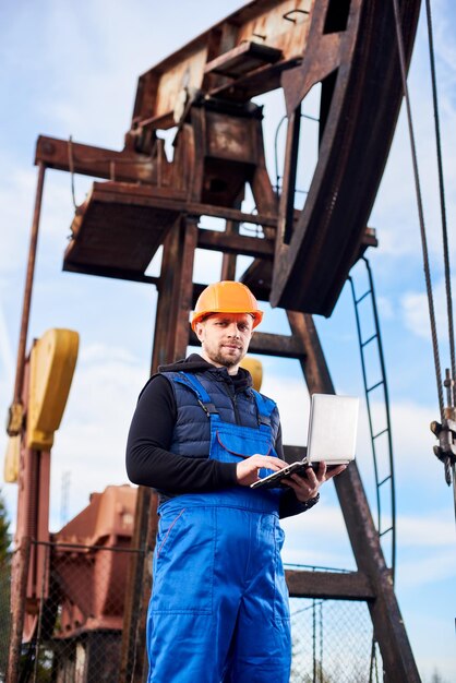 Oil engineer with a laptop standing next to an oil rig making notes in his computer