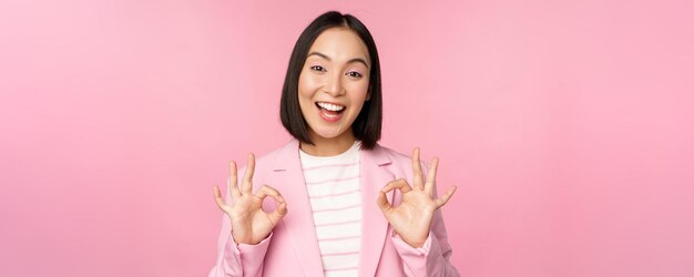 Okay excellent Businesswoman in corporate suit showing ok approval gesture recommending smth give positive feedback and smiling pleased posing over pink background