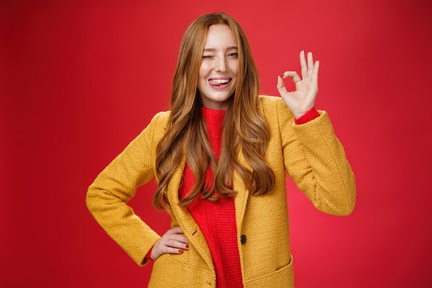 Okay got it. Portrait of carefree happy and confident good-looking charismatic ginger woman with freckles winking and showing tongue joyfully showing ok gesture, liking excellent work over red wall.