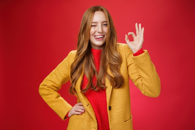 Okay got it. Portrait of carefree happy and confident good-looking charismatic ginger woman with freckles winking and showing tongue joyfully showing ok gesture, liking excellent work over red wall.