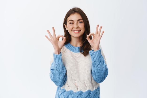 Ok, va bene. sorridente ragazza carina che mostra il segno ok e sembra soddisfatta, approva qualcosa di buono, loda un buon lavoro, in piedi con un maglione su bianco