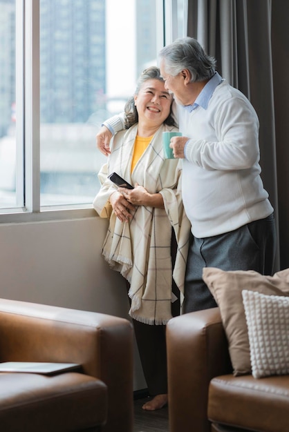 Vecchia coppia senior asiatica in pensione in piedi tenere la mano bevanda calda guardare fuori dalla finestra casa isolare conceptold senior in attesa di blocco è sopra lo sfondo interno della casa