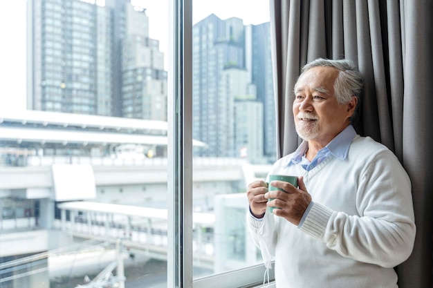 Free photo old asian senior retired age standing hand hold hot drink look outside window home isolate conceptold man waiting for lockdown is over home interior background