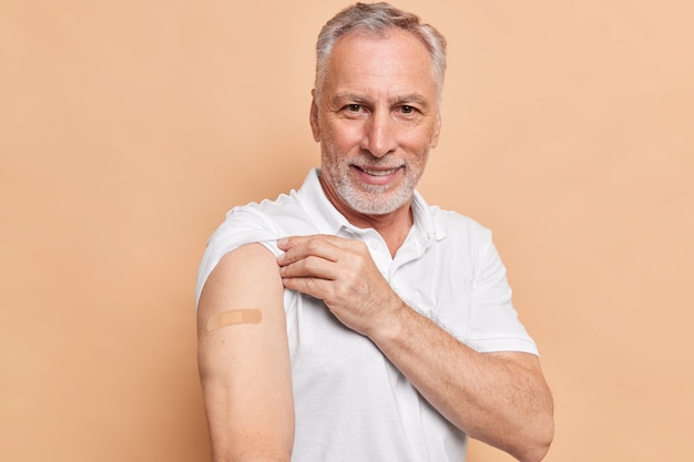 コロナウイルスの予防接種を受けたひげを生やした老人は、絆創膏で腕が茶色の壁に対するパンデミックのポーズ中に健康を気にかけていることを示しています