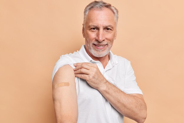 Old bearded man got vaccinated against coronavirus shows arms with adhesive plaster cares about healthy during pandemic poses against brown wall