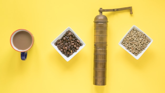 Бесплатное фото Старая кофемолка; сырые и жареные кофейные зерна и чашка кофе на ярко-желтом фоне