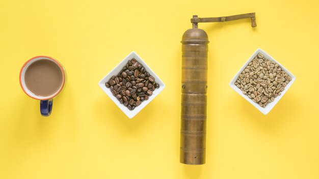 Old coffee grinder; raw and roasted coffee beans and coffee cup over bright yellow background