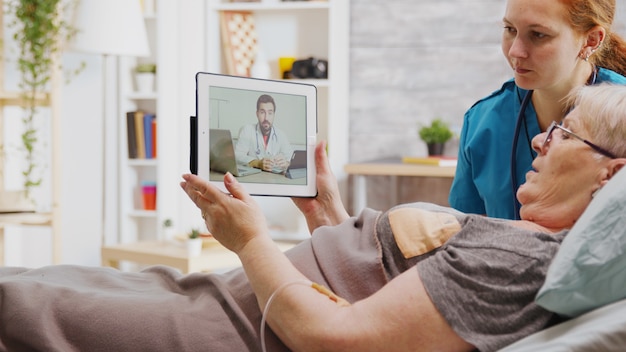 Old disabled woman lying in hospital bed having an online video call with a doctor. A nurse is next to her